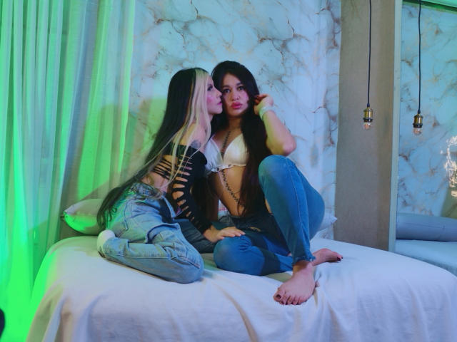 AbbyNdYliria, com cabelos longos, pretos e loiros, vestindo um sutiã branco e jeans rasgados, beija AbbyNdYliria, com cabelos longos, pretos, vestindo um sutiã bege e jeans azuis..