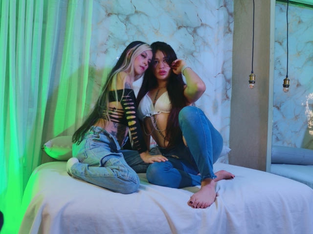 AbbyNdYliria, com cabelos longos, pretos e loiros, vestindo uma blusa listrada preta e amarela e jeans, está sentada em uma cama ao lado de AbbyNdYliria, com cabelos longos e escuros, um sutiã branco e jeans..