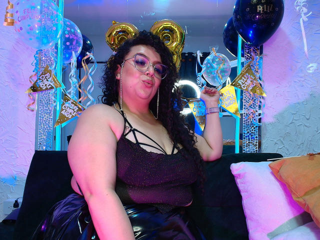 NatashaAmbeer with curly black hair, glasses, and pink lipstick, wearing a sparkly black top and shiny black pants, holding a clear balloon, pouting, on a black couch.