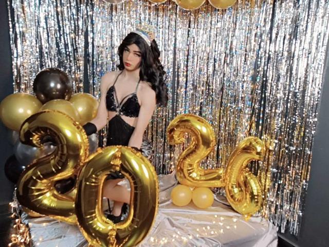 KendallSky with black lace bra, shiny black dress, and long wavy hair, sitting on table with gold "26" balloons and gold balloons, silver streamers in background.