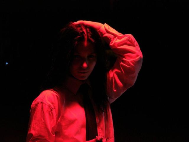 NetiShaw with dark curly hair, wearing a red-lit, puffy shirt, has one hand in her hair, looking slightly to the side with a serious expression.