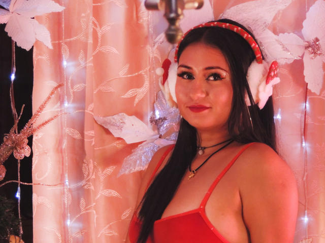 LunaSwt with long black hair, red dress, and red headphones, smiling with red lipstick, wearing a red headband, standing in front of pink and white fairy lights.