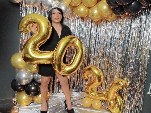CharlotteSweetX stands in black dress, holding gold "20" balloons, with a smaller gold "2" on the floor, surrounded by gold and black balloons, and silver streamers.