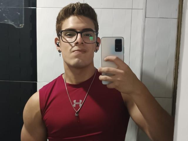 AngelEvil with glasses, brown hair, and a red sleeveless shirt, holding a phone, wearing earbuds, and a necklace, taking a mirror selfie showing a muscular upper body.
