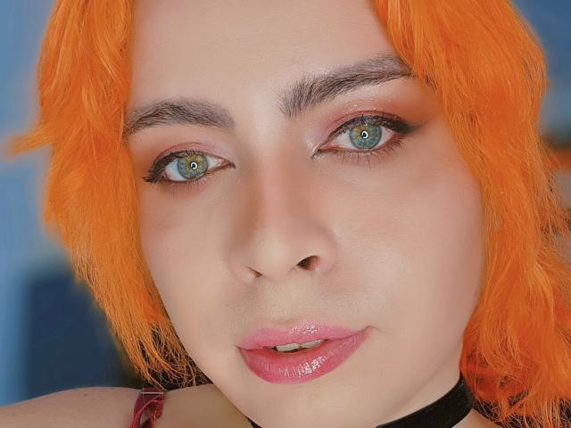 Close-up photo of TsFoxCandy with vibrant orange hair, pale skin, and blue eyes. TsFoxCandy has subtle makeup with pink eyeshadow and eyeliner, dark eyebrows, and glossy pink lipstick. TsFoxCandy's eyes are centered and slightly upward, with a neutral expression. The hair is slightly wavy and falls around the face. TsFoxCandy wears a black choker. The lighting is even, highlighting the skin's smooth texture and the bright orange hair. The image focuses on TsFoxCandy's face, with the blue background blurred. The overall composition is centered and symmetrical.
