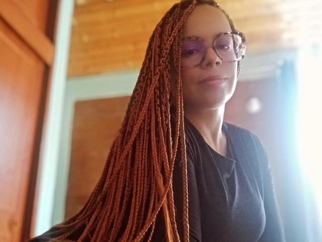 ElennaCruz with long, red-brown braids, wearing purple-tinted glasses and a black shirt, smiling slightly, wooden ceiling and door in background.