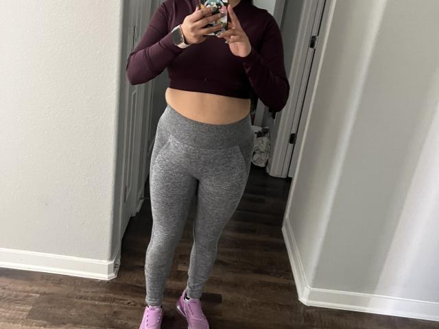 Rosily's midriff is exposed in a maroon crop top, wearing tight gray leggings and pink sneakers, standing in a doorway, holding a phone with a colorful case.