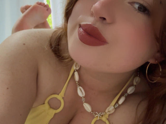 VicluX con rossetto rosso intenso, top giallo a halter, collana di conchiglie bianche e orecchini a cerchio dorati, che mostra le spalle scoperte e parte dei glutei sullo sfondo..
