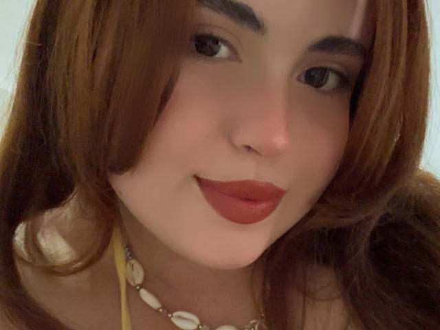 Close-up of VicluX with fair skin and brown, shoulder-length hair. VicluX has dark eyebrows, black eyes, and red lipstick. They wear a yellow necklace with white seashells. The image focuses on VicluX's face, which is slightly tilted to the right. The lighting is soft, highlighting VicluX's smooth skin and subtle blush. VicluX's expression is neutral, with a slight, almost closed smile. The overall composition is centered and tight, drawing attention to VicluX's facial features.