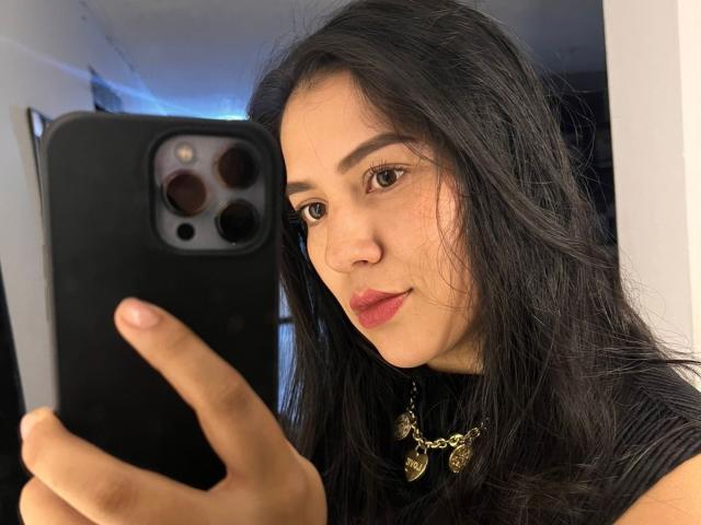 Velka with long black hair, brown eyes, and red lipstick, taking a selfie with a black phone, wearing a gold leaf necklace, showing a slight smile, holding the phone up.