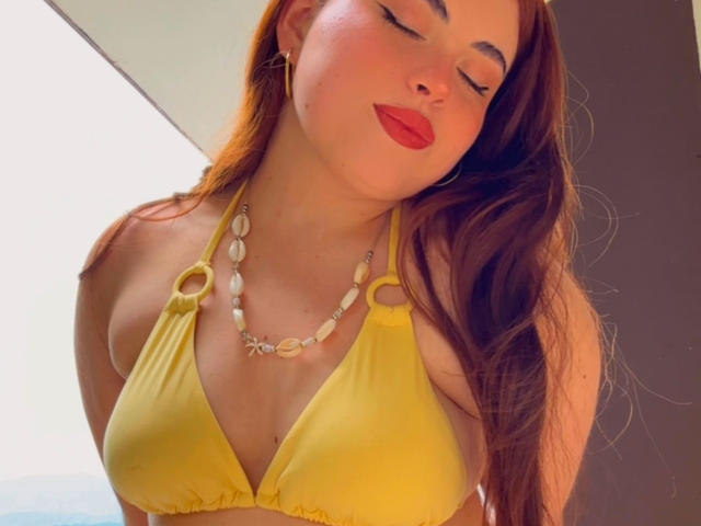 VicluX, les yeux fermés, porte un haut de maillot de bain jaune, du rouge à lèvres et un collier de coquillages, et ses longs cheveux bruns flottent. Ses seins sont de taille petite à moyenne..