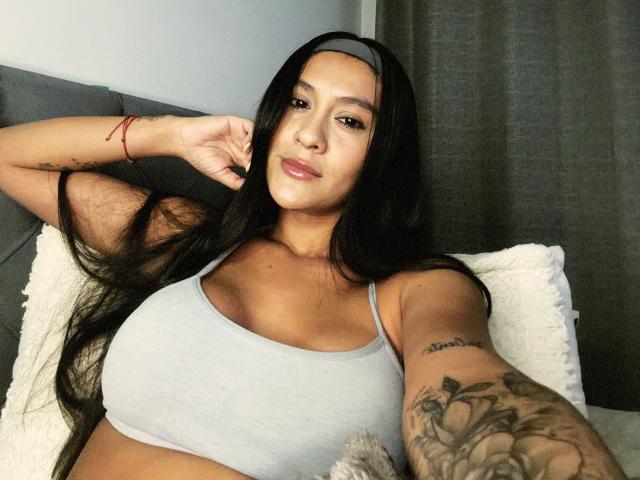 ThaliaAres with long black hair, gray headband, gray tank top, showing her big tits, tattooed arm, and red bracelet, lying on a gray couch with a white pillow.
