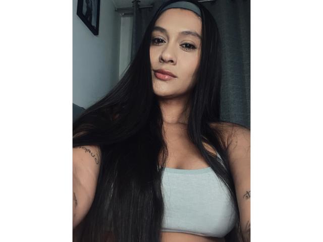 ThaliaAres ha lunghi capelli neri, indossa un reggiseno sportivo grigio, che mette in mostra piccoli seni, tatuaggi sulle braccia e un'espressione neutra. Indossa una fascia per capelli grigia..