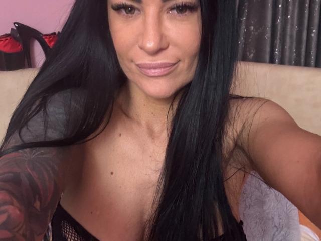 AndraD with long black hair, tan skin, and a tattoo on her right arm, smiling at the camera, wearing a black mesh top, showing her small tits.