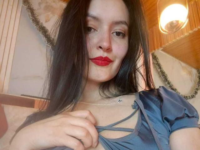 A photograph of AriaBonnet with long black hair, red lipstick, and light skin. She wears a blue dress with a crisscross neckline and a small heart necklace. Her right hand pulls at the dress, revealing more skin. The wooden ceiling and hanging lights are in the background. The composition is close-up, focusing on her face and upper body. The lighting is warm, highlighting her facial features. The texture of the dress is shiny and smooth. AriaBonnet's expression is slightly confident with a subtle smile.