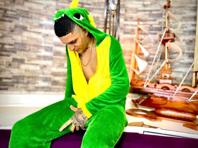 FodexKing in a green and yellow frog costume, sitting with head down, right hand touching his crotch, tattoos visible on left hand. Ship model in background.