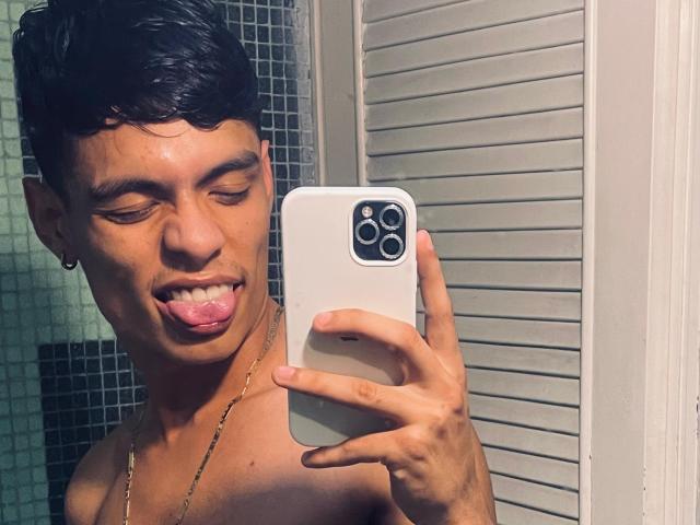 DaironLee with short black hair, tan skin, tongue out, shirtless, holding a white iPhone, gold chain, in a mirror selfie, wearing a small hoop earring.