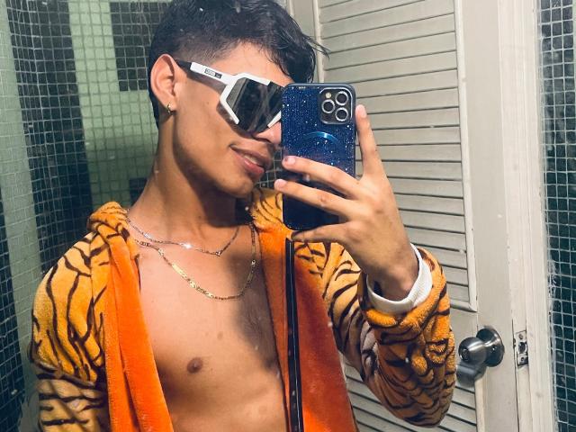 DaironLee with short dark hair, wearing tiger-striped hoodie, orange towel, white hexagonal sunglasses, gold chain, holding blue speckled phone, smiling, shirtless, showing toned abs, in bathroom.
