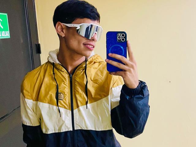 DaironLee with short black hair, wearing reflective sunglasses, gold and white jacket, holding blue phone, taking mirror selfie. His ass is not visible. He's smiling slightly.