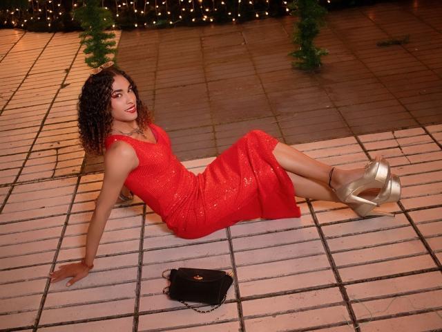 StefannyMia with curly brown hair, red sparkly dress, and silver heels, sitting on a white-tiled floor, legs extended, black purse on ground, smiling, with warm lighting.