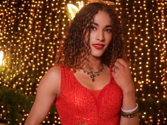 StefannyMia with curly brown hair, red sequined dress, red lipstick, necklace, and bracelets, smiling, holding her dress, glowing yellow fairy lights in the background.