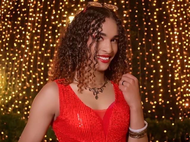 StefannyMia with curly brown hair, red sequined dress, smiling, wearing a necklace and bracelets, holding her hair, glowing boobs, red lipstick, and gold sunglasses on her head.