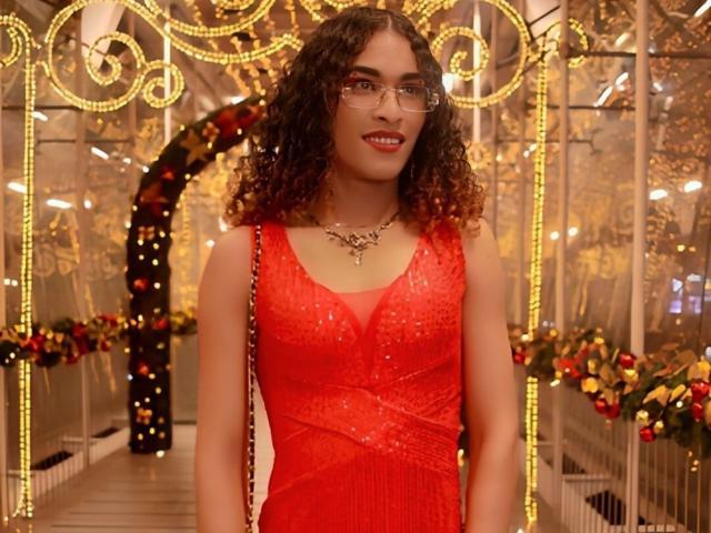 StefannyMia with curly brown hair, glasses, red dress with sparkles, necklace, smiling, medium tits, cute, standing in a warmly lit, ornate, decorated hallway.