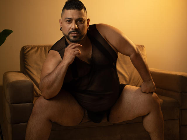 JacobWoods, a chubby, muscular man with dark skin and short hair, sits on a beige couch in black sheer underwear, legs spread, hand near chin, looking serious.