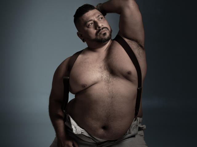 Shirtless JacobWoods with a hairy chest, wearing suspenders and gray pants, has a chubby belly, raised arm, and a confident, slightly smug expression. His balls are partially visible.