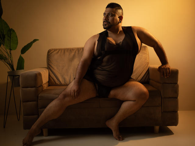 JacobWoods, a chubby man with dark hair and beard, sits on a beige sofa wearing a black sheer tank top and black underwear, legs spread, arms resting on sofa.