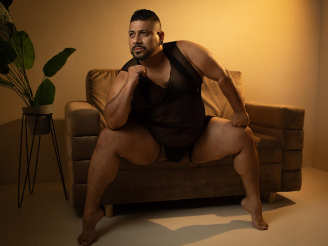 JacobWoods, a muscular, hairy man with a beard, wearing a sheer black tank top and black underwear, sits on a brown sofa, legs spread, gripping the top with his right hand.