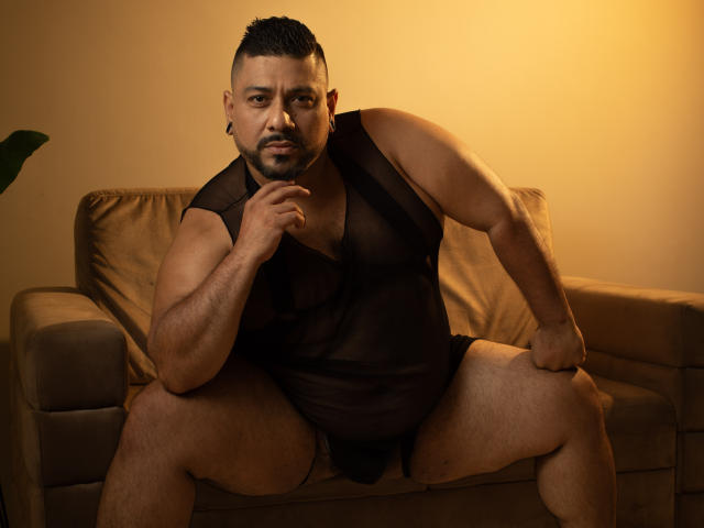 JacobWoods, a muscular, dark-skinned man with a beard, sits on a brown sofa, wearing a sheer black mesh shirt, exposing his chest and black underwear, legs spread.