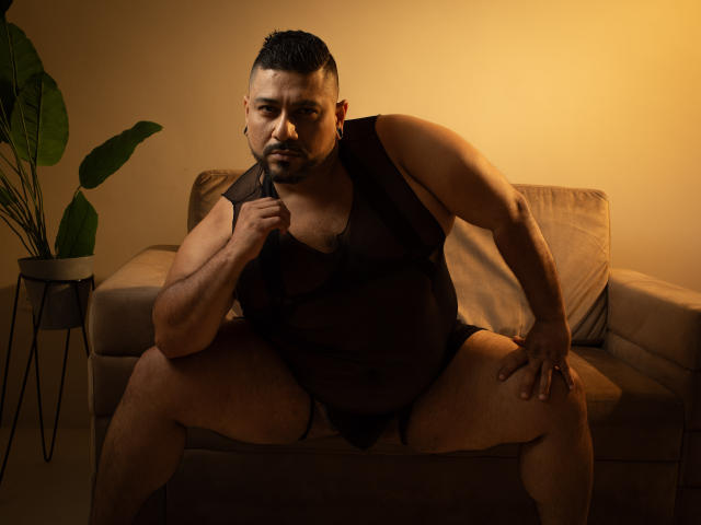 JacobWoods, a chubby man with short black hair, wearing a sheer black mesh tank and black underwear, sits on a beige couch, legs spread, holding the tank with a serious look.