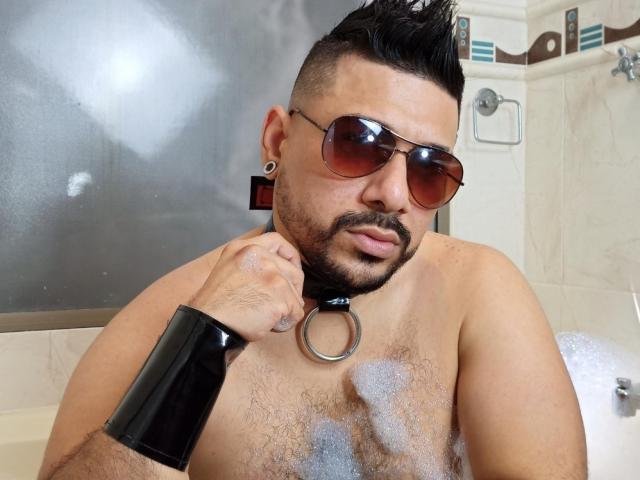 Shirtless JacobWoods with dark hair, beard, and spiked style, wearing brown sunglasses, black wrist cuff, and collar with ring, in a bubble-filled bathtub, looking serious.