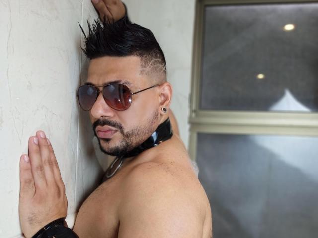 shirtless JacobWoods with spiked black hair, brown aviator sunglasses, black collar, and ear studs, standing against a wall, hands up, looking back with a serious expression.