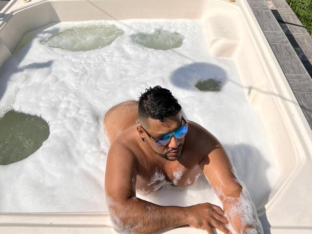 JacobWoods, naked, in a bubbling hot tub, leaning forward with soapy water covering his chest and arms, wearing blue-tinted sunglasses, and looking up with a serious expression.