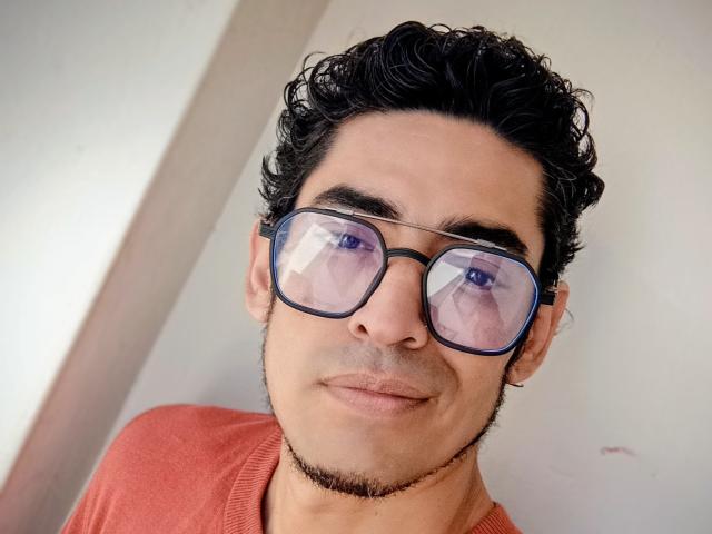 Rahullooo with curly black hair, wearing blue glasses reflecting his face, has a light beard and is smiling slightly, dressed in a red shirt.