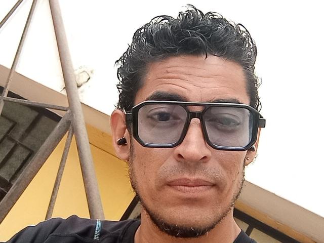 Rahullooo has curly black hair, black glasses, a small goatee, and black ear studs. He's wearing a black shirt with a zipper. His nose is slightly upturned.