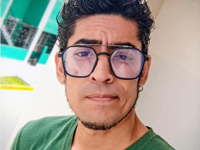 Rahullooo with curly black hair, green shirt, and black glasses, has a serious expression and a light stubble, showing purple-tinted lenses. Medium skin, slightly goofy face.
