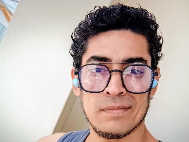Rahullooo with curly black hair, blue earbuds, and black-framed glasses reflecting his face, wearing a gray sleeveless shirt, has a light brown beard and a neutral expression.