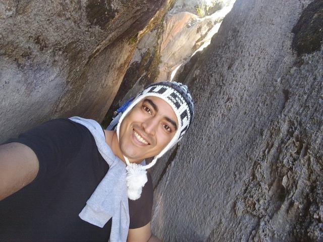 Rahullooo with dark brown eyes, smiling, wearing a black shirt, gray scarf, and white-and-black striped knit hat, standing between rocky walls.