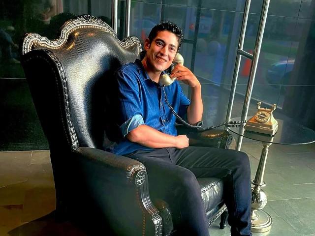 Rahullooo sits in a black, ornate leather chair, wearing a blue shirt and black pants, holding a phone to his ear, smiling, with a vintage phone on a stand.