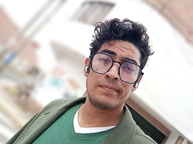 Rahullooo with curly black hair, glasses, green shirt, white V-neck, green jacket, ear gauges, slight smile, and blurred background. Medium skin, slight stubble, looking down at camera.