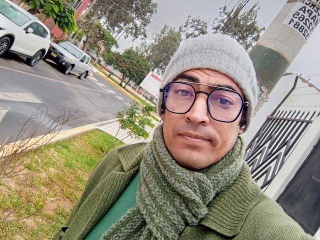 Rahullooo with glasses, gray beanie, green scarf, and green coat, standing outside, looking at the camera, slightly smiling. His face has a light skin tone.