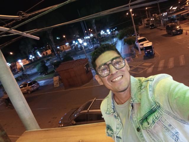 Rahullooo with green eyes, short black hair, and glasses, wearing a green and white denim jacket, smiling at the camera, standing in a nighttime urban setting.