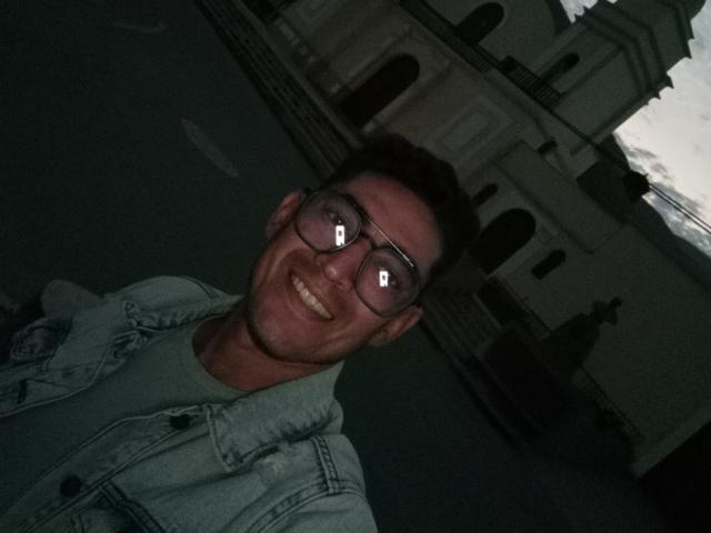 Rahullooo, smiling with glasses reflecting lights, wearing a greenish denim jacket, dark short hair, and a light green shirt, standing in a dimly lit outdoor area.