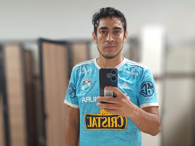 Rahullooo, with dark curly hair and brown skin, wearing a blue sports jersey with "Jatari" logo, holding a black phone, taking a selfie in a locker room.