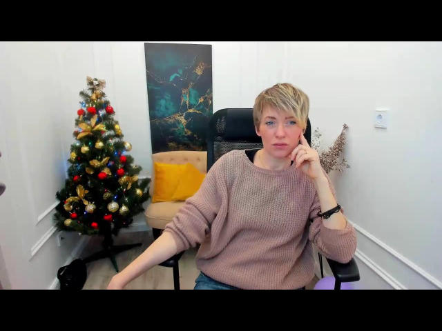 AlisArinti with short blonde hair, wearing a pink knit sweater, sitting in a black chair, talking on a cellphone, next to a decorated Christmas tree and a wall painting.