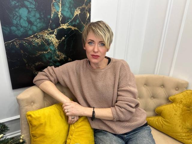 AlisArinti with short blonde hair, pink sweater, and blue jeans, leaning on a beige couch with yellow pillows, looking slightly to the side.