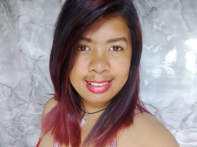 MariahSquirting69 has medium brown skin, straight dark red and black hair, pink lipstick, and a black choker. She's smiling with pink teeth, wearing a red top.